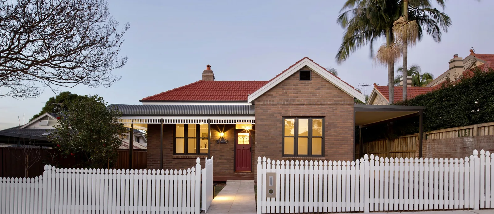 A weatherboard and brick home with a white picket fence, lit warmly at dusk